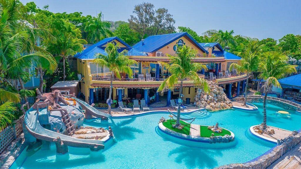 Complejo turístico con piscina grande, tobogán curvado, palmeras, casa de techo azul y varias zonas de descanso al aire libre