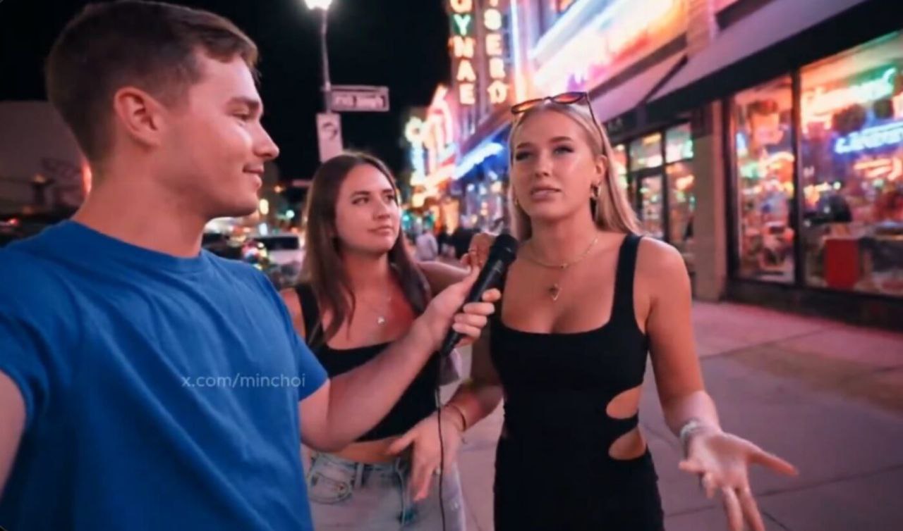 Entrevistador sostiene micrófono mientras conversa con dos mujeres en una calle iluminada por coloridos letreros de neón durante la noche.