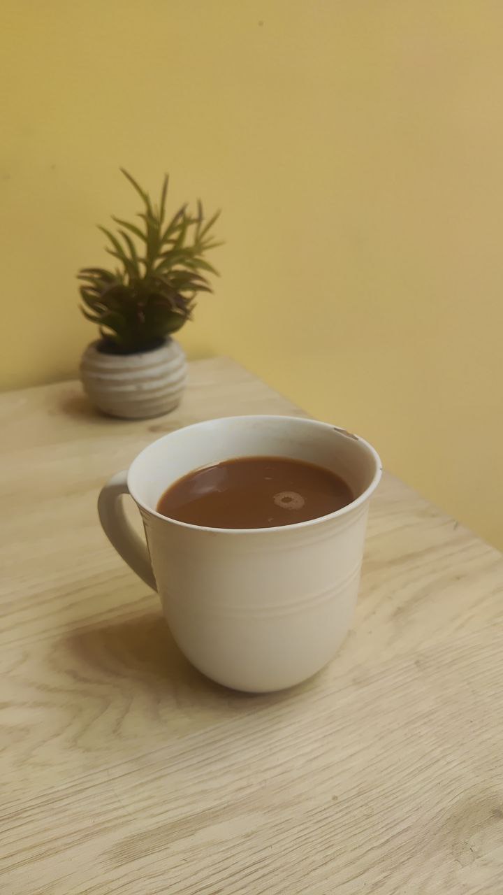Taza blanca con bebida caliente sobre mesa de madera clara, acompañada de una pequeña planta decorativa al fondo
