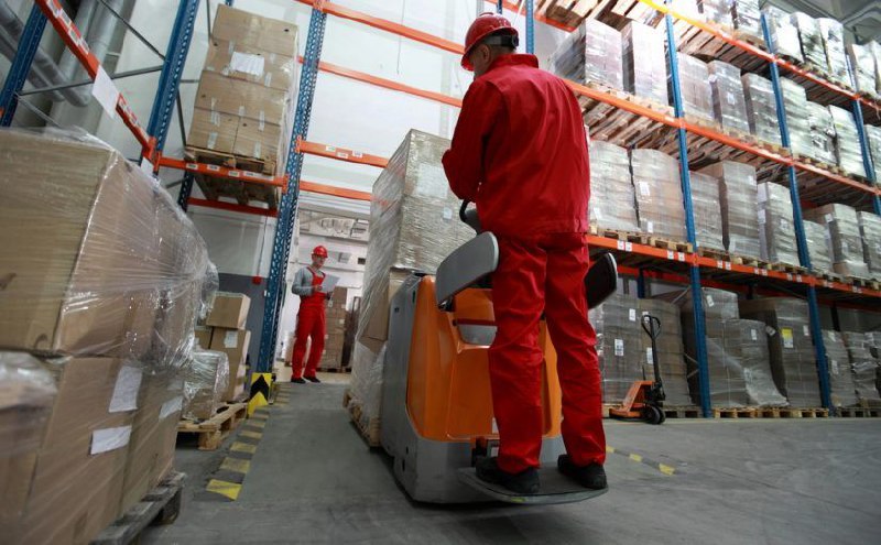 Dos trabajadores con uniforme rojo operan maquinaria en un almacén lleno de estanterías con cajas y palets apilados.