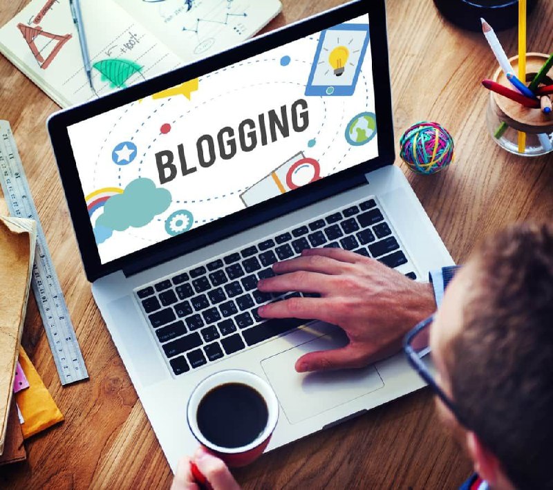 Persona usando una laptop que muestra la palabra 'BLOGGING', rodeado de elementos gráficos coloridos y una taza de café.